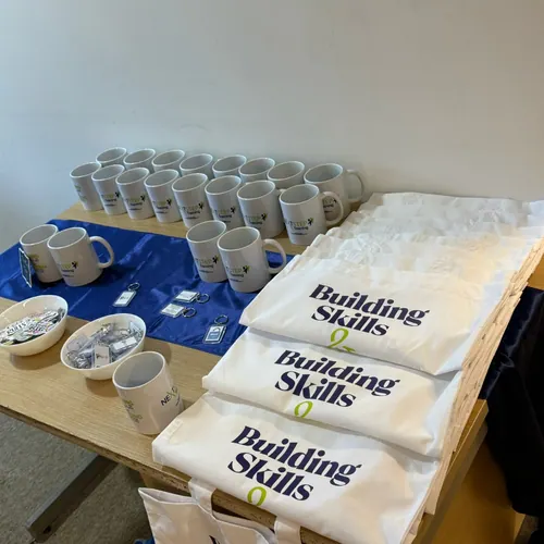 Building Skills tote bags and mugs are arranged on a table.